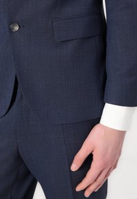 Navy blue suit jacket featuring a textured fabric, single button closure, and a white shirt cuff visible beneath the sleeve with smooth texture.