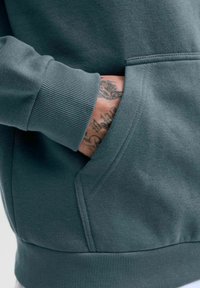 Dunkelgrüner Sweatshirt mit gerippten Bündchen und einer Fronttasche, aus einem glatten Baumwollstoff mit subtilen Steppnähten.