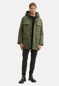 Olivovo zelená parková bunda s kapucňou, viacerými prednými vreckami, zipsom a štruktúrovanou textíliou, spárovaná s tmavými nohavicami a čiernymi čižmami.