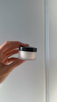 Hand holding a small frosted jar with a black lid labeled "Laura Mercier" against a plain light gray background.