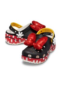 Schwarze und rote Clogs mit einem Polka-Dot-Muster, ausgestattet mit großen, roten, geknüpften Pailletten-Schleifen und weißen Blumenakzenten am Riemen. Texturiertes und leichtes Design.