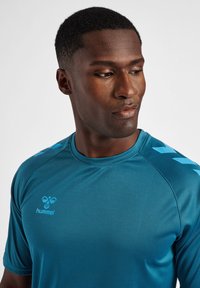 Turkos atletisk T-shirt tillverkad av slätt, fuktavvisande tyg. Har en rund halsringning, korta ärmar och blå chevronaccenter på axlarna.