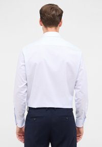 Uomo che indossa una camicia a maniche lunghe a quadretti blu chiaro infilata in pantaloni formali blu scuro, visto da dietro su uno sfondo semplice.