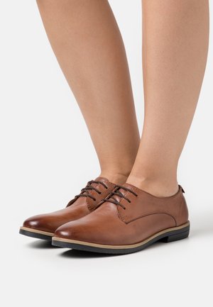 Chaussures en cuir marron à lacets avec un bout arrondi, des coutures contrastantes et une semelle en caoutchouc noir bas. Texture lisse avec des accents minimaux.
