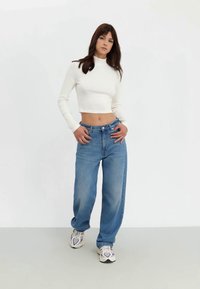 Top cropped de manga longa branca combinado com jeans leves de corte solto azuis. A modelo usa tênis cinzas. Fundo simples com tons neutros.