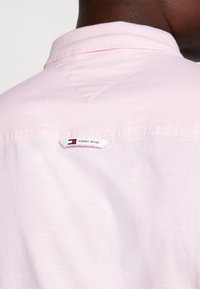 Nahaufnahme einer Person, die ein hellrosa Hemd trägt, auf dem im oberen Rückenbereich ein weißes Tommy Jeans Etikett angenäht ist.