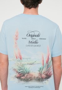 Ανοιχτό μπλε βαμβακερό μπλουζάκι με γραφικό σχέδιο από ροζ χόρτα και λίμνη στο πίσω μέρος, με τυπωμένο κείμενο και μαλακή υφή.