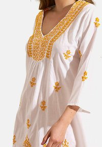 Tunique blanche avec broderie florale jaune. Design en V, manches longues, tissu léger, avec des détails intricats le long de l'encolure et de l'ourlet.