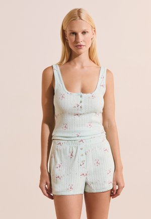Mujer rubia con camiseta de tirantes azul claro con canalé y pantalones cortos a juego con pequeño estampado floral rosa y detalles de botones, de pie contra una pared beige.