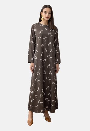 Donna che indossa un lungo vestito marrone con motivi floreali bianchi, maniche lunghe, scarpe marroni a punta e grandi orecchini dorati, in piedi su uno sfondo semplice.