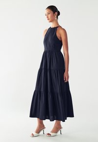 Žena v bezrukávových námorníckych šatách maxi s tierovaným strihom, oblečená v bielych sandáloch s otvorenou špičkou a perlovými náušnicami, stojí proti jednoduchému pozadiu.