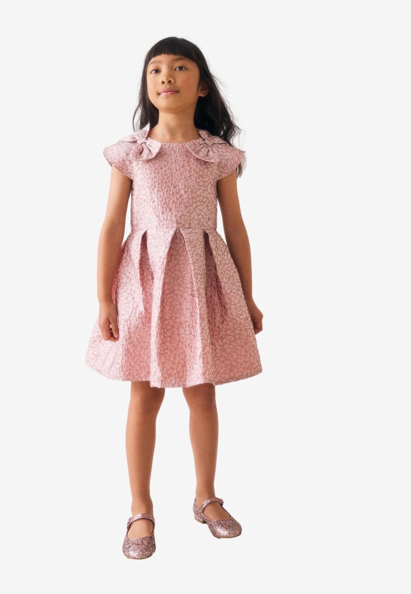 Roosa varrukateta kleit tekstuurse kangaga, lehvikukujulise seeliku ja vibukujundusega õlgadel. Kaunistatud läikivate ja sädelevate kingadega.