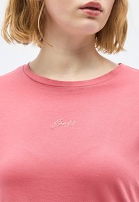 Γυναίκα με κοντά ξανθά μαλλιά που φοράει ροζ πουκάμισο με το κείμενο «Boss» κεντημένο με άσπρα γράμματα στο στήθος.