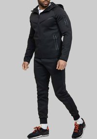 Veste zippée noire avec capuche, poches latérales zippées et pantalon de survêtement. Tenue athlétique décontractée au design ajusté et à tissu texturé.