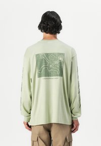 T-shirt à manches longues en coton vert clair, avec un imprimé graphique au dos et des motifs détaillés le long des manches.