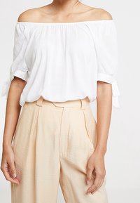 Blusa bianca off-the-shoulder con maniche a sbuffo e dettagli annodati; indossata sopra pantaloni beige a vita alta con pieghe e tessuto testurizzato.