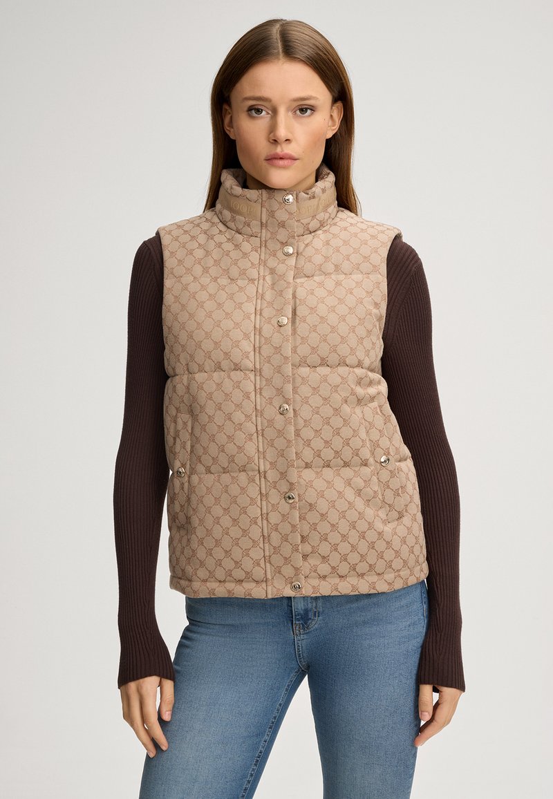 Beige gesteppte Weste mit hohem Kragen, die ein dezentes Musterdesign und dekorative Knöpfe aufweist, kombiniert mit einem dunkelblauen Langarm-Oberteil.