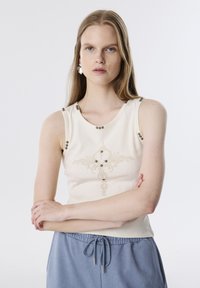 Mouwloze crèmekleurige top met decoratieve borduursels en metalen accenten; aansluitend ontwerp met een ronde halslijn. Gepaard met lichtblauwe broek met trekkoord.