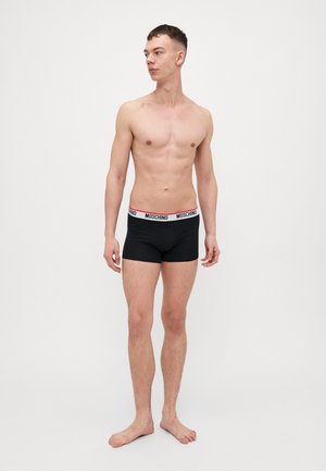 Crne bokserice s bijelim i crvenim pojasom "MOSCHINO". Tkanina izgleda glatko s pamučnom teksturom. Model je pozirao opušteno.