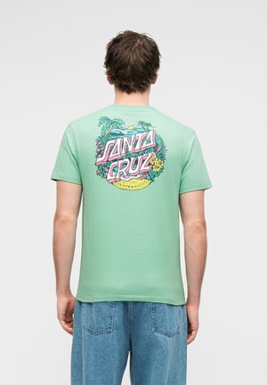 Mann trägt ein mintgrünes T-Shirt mit einer bunten "Santa Cruz Skateboards"-Grafik auf dem Rücken, die tropische Pflanzen und einen Sonnenuntergang zeigt, kombiniert mit blauen Jeans.
