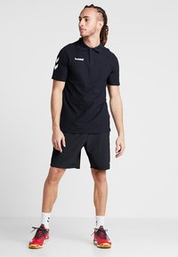 Zwarte poloshirt met korte mouwen, voorzien van een wit logo op de borst en een chevronpatroon op de mouwen. Geko Zen met zwarte shorts en rode sportschoenen.