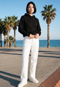 Giovane donna con felpa nera e pantaloni bianchi in piedi su un'area lastricata vicino a palme con l'oceano sullo sfondo in una giornata limpida.