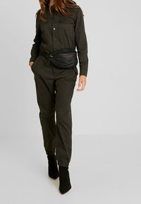 Mono de utilidad verde oscuro con mangas largas y dos bolsillos en el pecho, combinado con una riñonera negra y botas negras hasta el tobillo.
