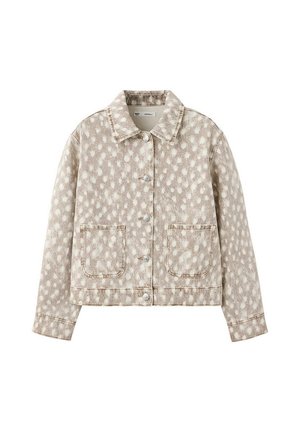 Joven mujer con chaqueta vaquera beige con manchas blancas y pantalones a juego, de pie contra una pared lisa de color gris claro.