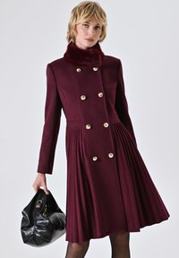 Bordeaux wollen jas met een faux fur kraag, dubbele goudkleurige knopen, geplooide rok en een zwarte tas met een gouden accent.