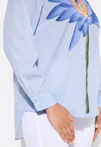 Blusa blu a righe con motivo floreale nelle tonalità di blu e giallo, con polsini bottonati e una fessura laterale. Realizzata in tessuto leggero.