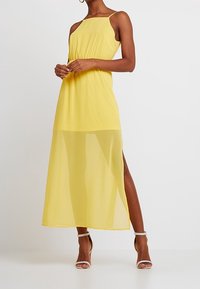 Robe longue jaune en tissu léger et transparent, dotée de fines bretelles, d'une taille cintrée et d'une fente latérale pour plus de fluidité.