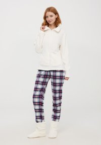 Giacca in pile bianca con zip e design a coste, abbinata a pantaloni del pigiama a quadri in navy, rosso e bianco, e pantofole morbide bianche.