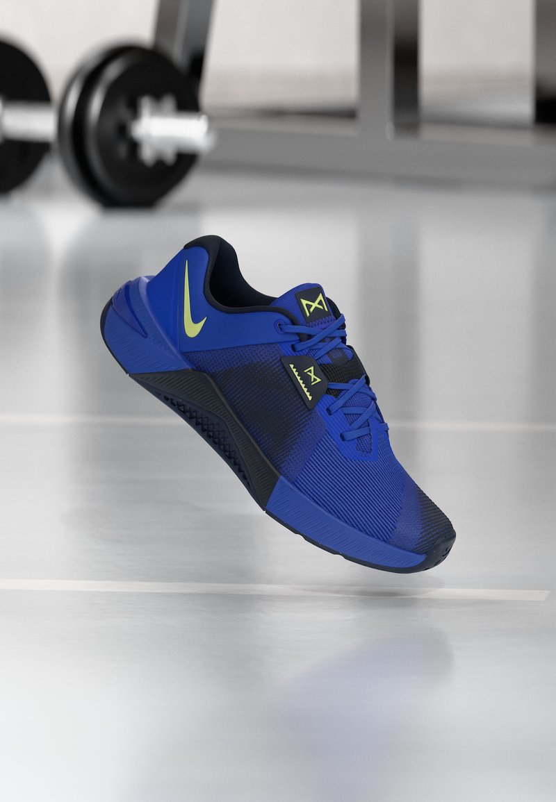 Μπλε και μαύρο αθλητικό παπούτσι Nike με λογότυπο σε χρώμα λάιμ, που αιωρείται πάνω από δάπεδο γυμναστηρίου με βάρη στο θολό φόντο.