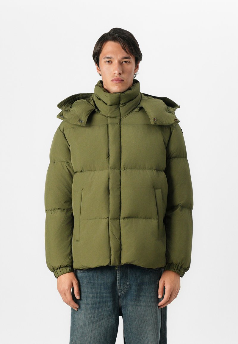 Hombre vestido con una chaqueta acolchada de color verde oliva con cuello alto y capucha desmontable, combinado con jeans azules, de pie contra un fondo blanco.