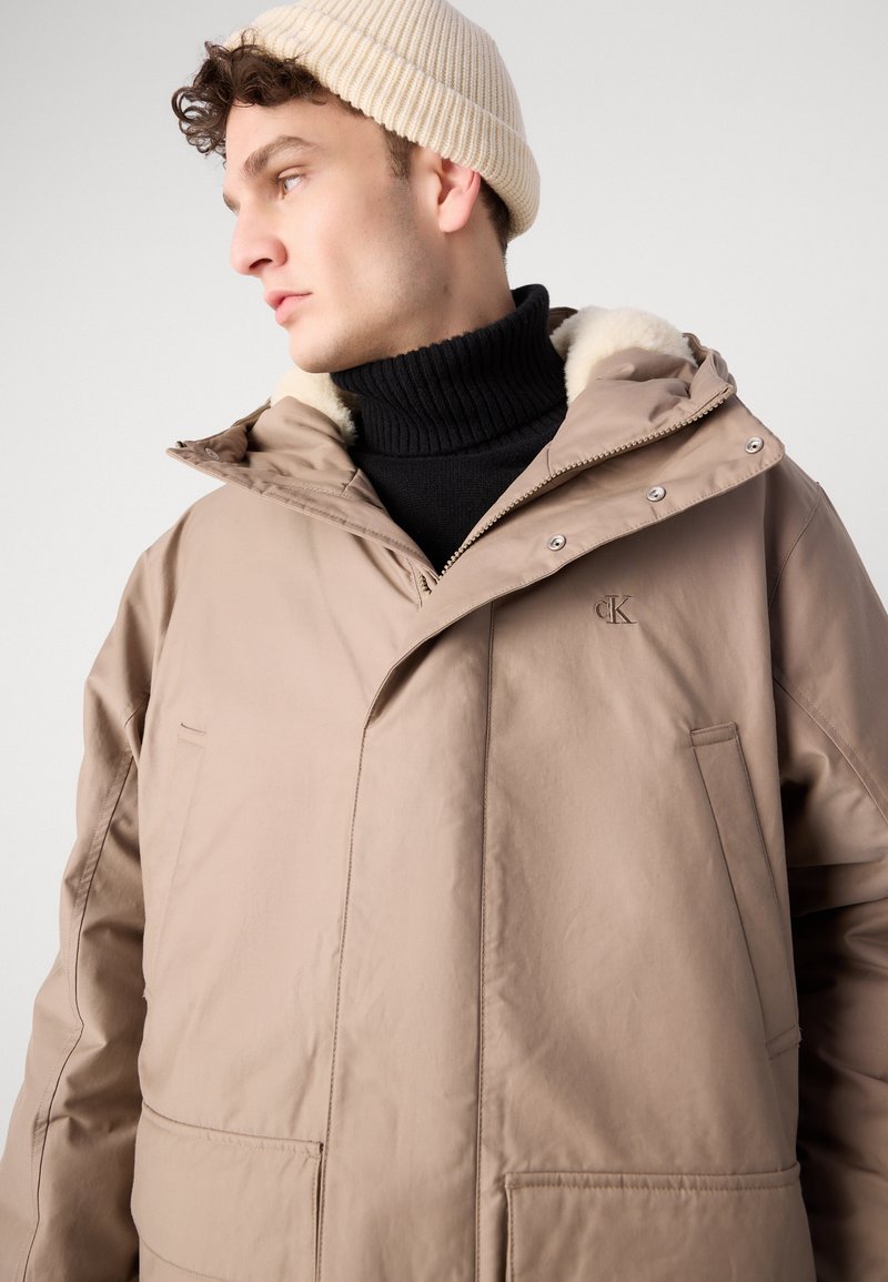 Beige geïsoleerde jas met een hoge kraag en bontvoering, voorzien van een rits aan de voorkant, twee zijzakken en het CK-logo op de borst. Gebreide beige muts.