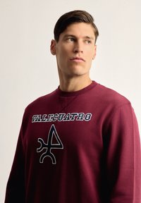 Sudadera burdeos con cuello redondo y puños de canalé. Presenta el texto "VALECUATRO" y un logotipo gráfico distintivo en negro. Textura suave.