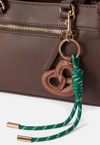 Hnedá kožená kabelka s zlatým zipsom, hnedým koženým srdcovým príveskom a zelenými pletenými ramienkami s kovovými koncami.
