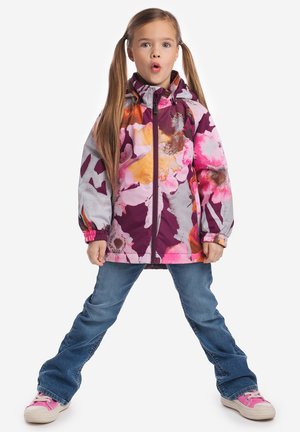 Jong meisje met lang haar in vlechtjes, gekleed in een kleurrijk bloemenjasje, blauwe spijkerbroek en roze sneakers, staand met gespreide benen en open mond.