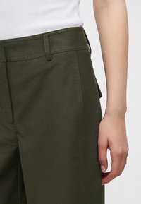 La main d'une femme repose le long du corps, elle porte un pantalon taille haute vert foncé et un haut blanc côtelé sans manches.