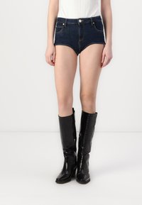 Temne modri jeans šorti, visoko pasu z sprednjimi žepi in podrobnostmi šivanja, v kombinaciji s črnimi, visokimi, sijajnimi čevlji iz krokodiljeve kože.
