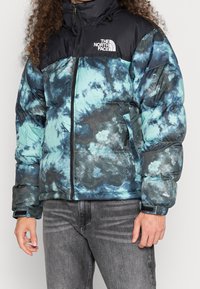 Donsjack met een blauw-zwarte tie-dye print, voorzien van een hoge kraag, ritssluiting en logo op de borst. Zachte, opgeblazen textuur.