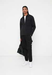 Črna zip-up jakna z žepom na prsih, v kompletu s cargo hlačami, teksturiranimi tkaninami in belo majico pod njo. Bele superge v roki.