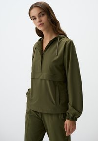 Veste légère à capuche, de couleur vert olive, avec une poche avant, un cordon de serrage réglable et des poignets élastiques ; texture lisse.