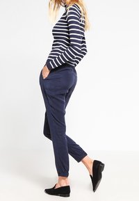 Pantalones estilo pijama de color azul marino con puños elásticos y bolsillos laterales, combinados con una blusa de rayas de mangas largas en azul marino y blanco. Zapatos negros de tipo slip-on.