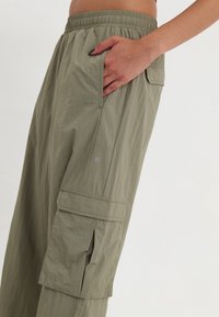 Lekkie spodnie cargo w stonowanej oliwkowej zieleni, z elastycznym pasem, bocznymi kieszeniami i fakturowanym materiałem o delikatnym połysku.