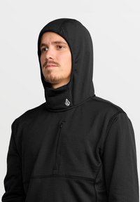 Haut de sweat à capuche noir avec un col montant et une capuche, fabriqué en tissu texturé. Il présente un petit logo sur le col et une poche poitrine zippée.