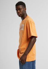 Orange bomulds-t-shirt med korte ærmer, har hvid skrift på fronten, afslappet pasform, rund halsudskæring og en glat tekstur.