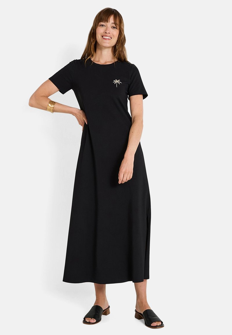 Robe maxi noire à manches courtes avec un col rond, ornée d'un embellissement de feuille de palmier sur la poitrine et de fentes sur les côtés. Portée avec des sandales noires.