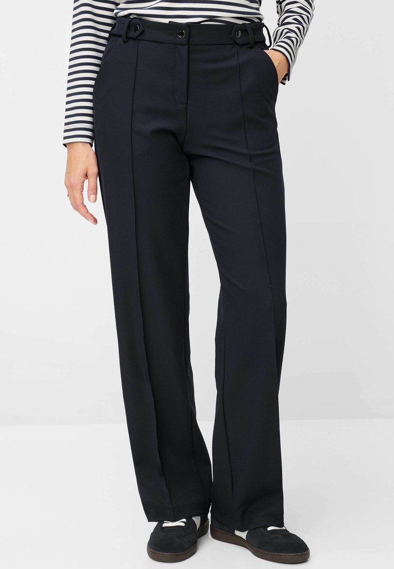 Personne portant un pantalon droit bleu marine avec des coutures avant, une chemise à manches longues rayée et des chaussures noires décontractées, debout les mains dans les poches.