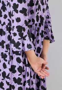 Vestido morado con patrón floral negro, con mangas acampanadas y cintura fruncida, hecho de una tela ligera. Detalles de costura visibles.
